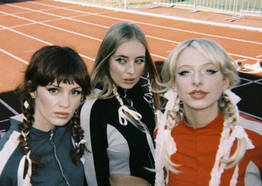 Three members of the band Blusher stood on an athletics track all with plaits and bows in their hairx